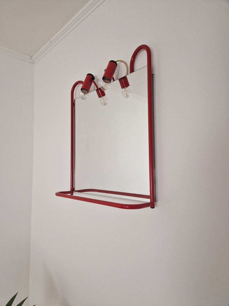 May include: A red metal framed mirror with a shelf and three light bulbs. The mirror is mounted on a white wall.