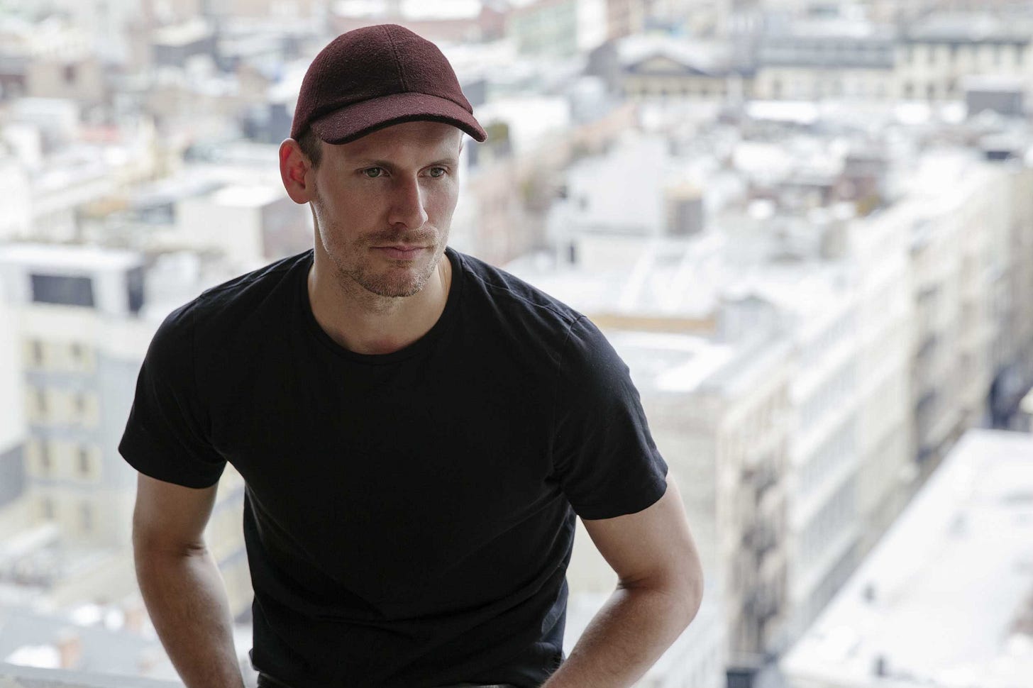Designer Dion Lee in a black t-shirt and a dark red cap, looking thoughtfully to his right. He is in front of a window with a soft-focus view of a city skyline in the background.