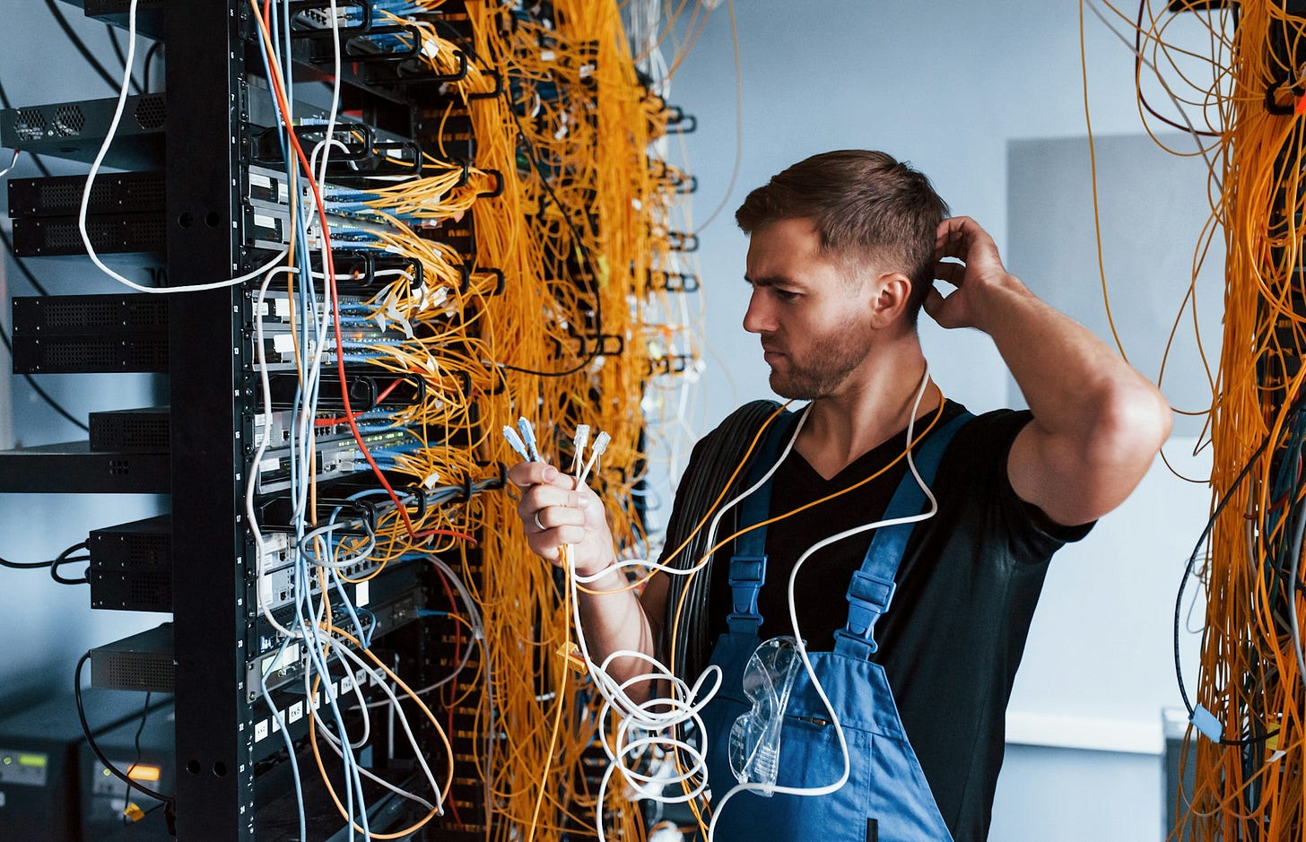 A man holds loose cables and scratches his head beside racks of network equipment densely packed with yellow and white wiring. A man holds loose cables and scratches his head beside racks of network equipment densely packed with yellow and white wiring.