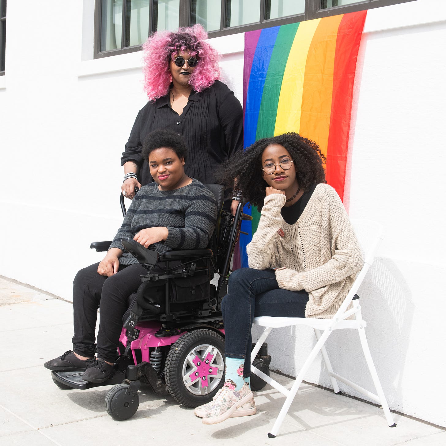 Three young Black women posing together against a rainbow Pride flag backdrop. One woman uses a hot pink motorized wheelchair, another stands behind wearing pink hair and sunglasses, and the third sits on a white chair wearing glasses and a cream cardigan. Photo credit: Disabled and Here.