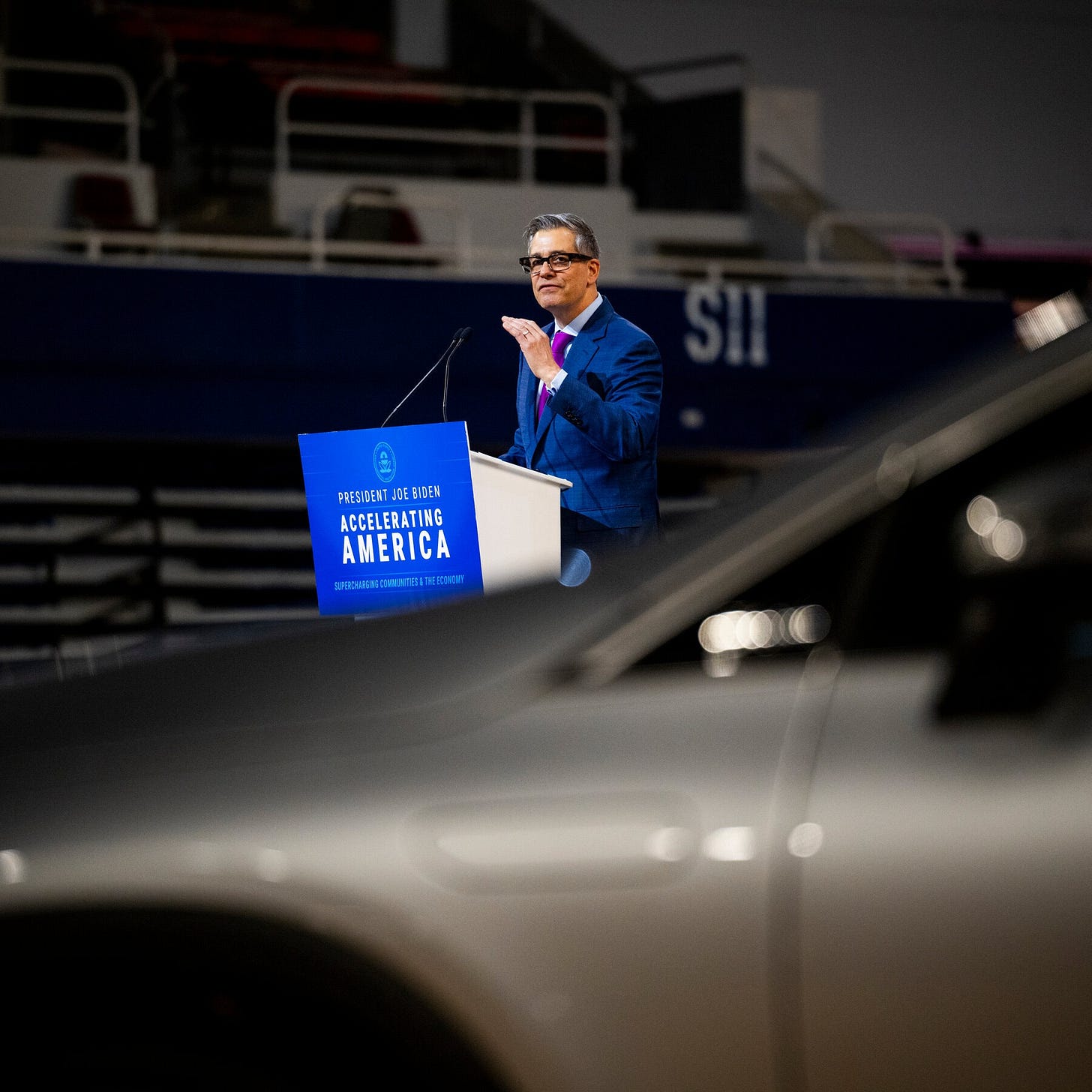 John Bozzella, dressed in a blue suit and red tie, speaks from a white podium. 