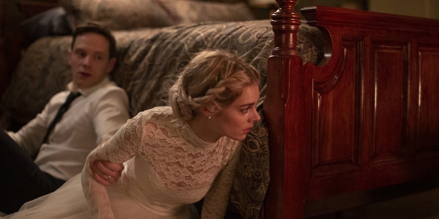 Alex and Grace Le Domas hiding behind a bed on the floor. Alex is grabbing Grace's arm as she peeks from around the corner of the bed frame. They're both in their wedding attire.