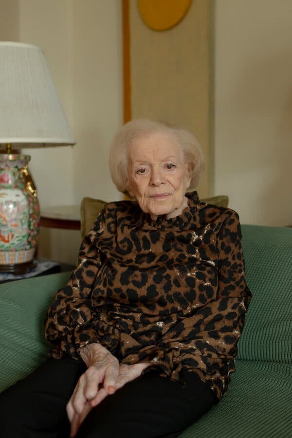 An older woman wearing a fancy animal print blouse and sitting on a green couch.