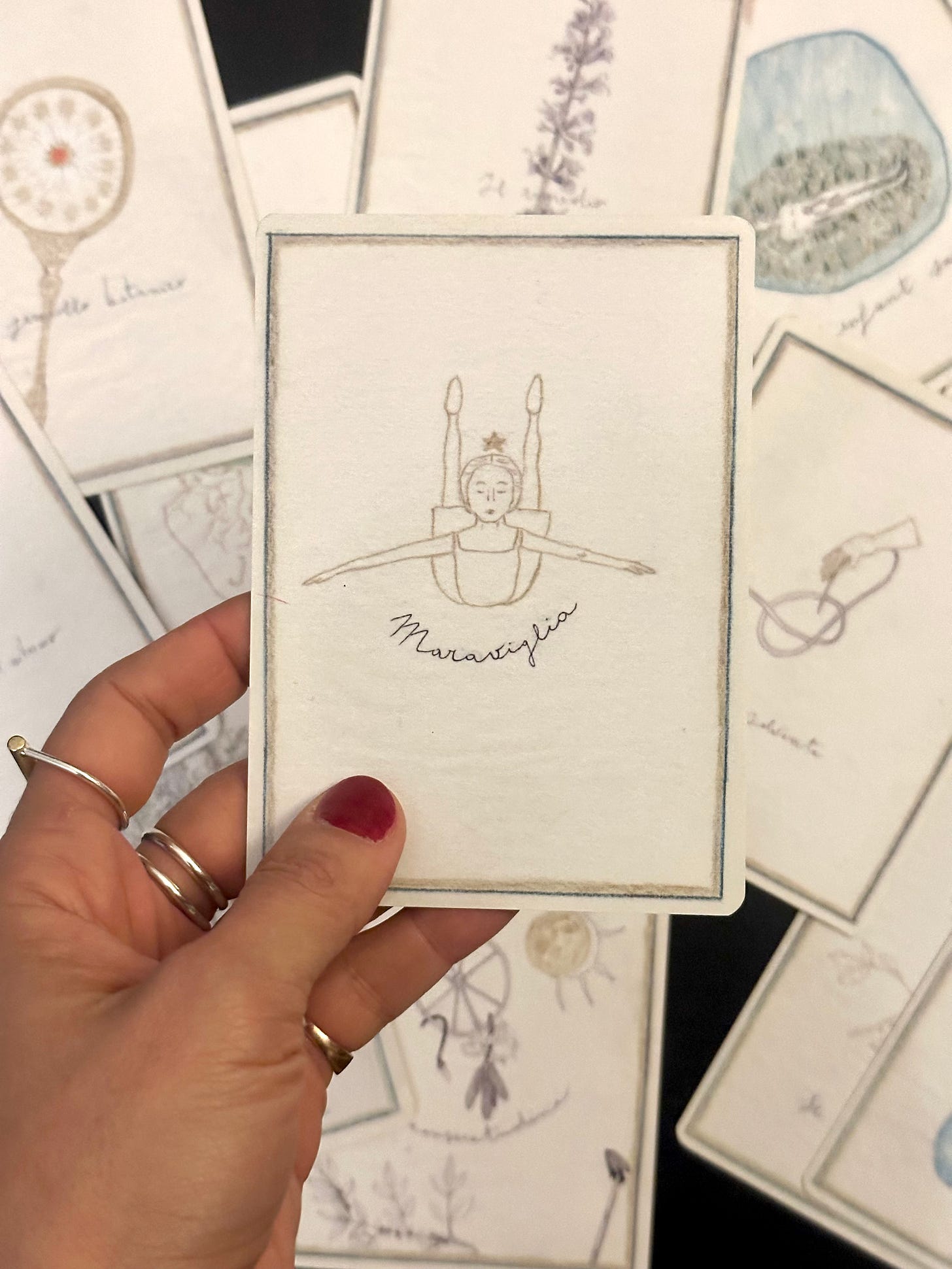 Hand holding a beautifully illustrated oracle card featuring a dancer in mid-motion, surrounded by other cards with delicate botanical and celestial drawings, showcasing an artistic, vintage-style collection
