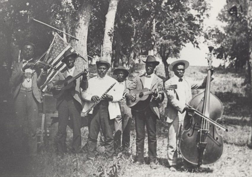 Historical black & white photo of seven Black men holding musical instruments (a violin, two guitars, a string bass and a clarinet) in an outdoor park in front of trees. One tree has an American flag attached to it. The men are dressed in suits and wearing a variety of broad-brimmed hats. The left side of the photo is smudged, partly obscuring the man with the fiddle. Historical black & white photo of seven Black men holding musical instruments (a violin, two guitars, a string bass and a clarinet) in an outdoor park in front of trees. One tree has an American flag attached to it. The men are dressed in suits and wearing a variety of broad-brimmed hats. The left side of the photo is smudged, partly obscuring the man with the fiddle.
