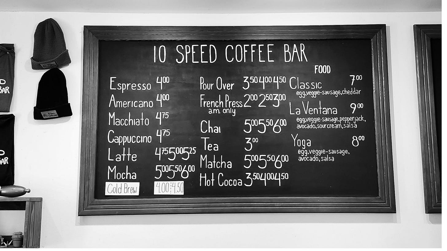A coffee shop menu written in white chalk on a black board is framed and hanging on a white wall.