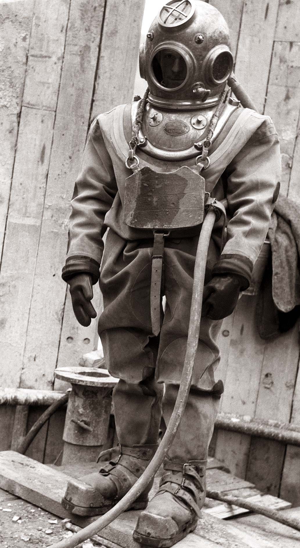 A historical black-and-white photograph of a diver wearing a heavy metal diving suit at the Ožbalt hydroelectric power plant construction site in 1958. The diver is equipped with a large helmet and weighted boots, standing in shallow water near a wooden structure.
