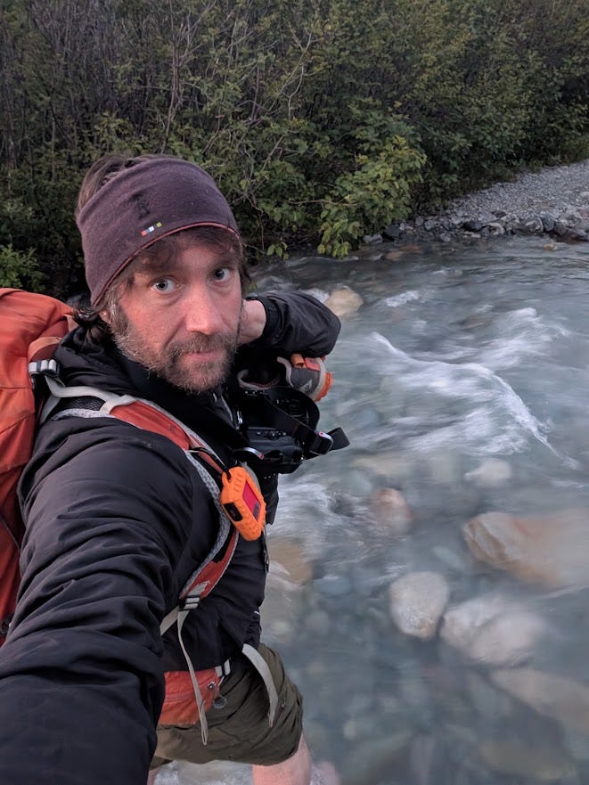 A dim selfie beside a milky, blue-grey glacial stream at twilight. A bearded man in a dark maroon headband and black jacket looks into the camera with tired, wide eyes, an orange pack on his back with a small orange GPS communicator clipped to the strap and a camera slung across his chest. Behind him, brushy green banks and a gravel bar line the rushing water. The light is the deep dusk of an Alaska summer night.