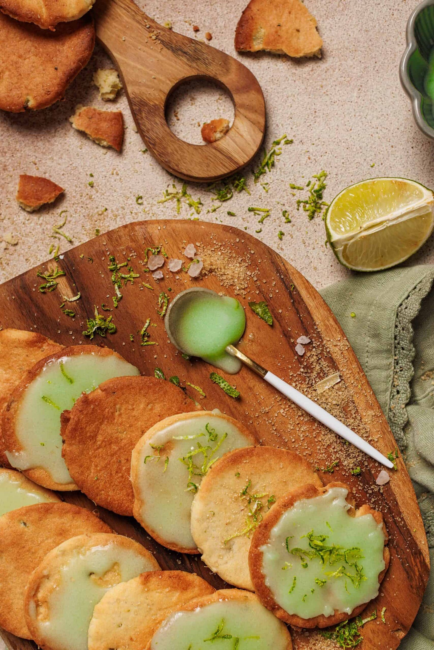 Lemongrass cookies and a spoon of green icing on a messy table with limes and a napkin.