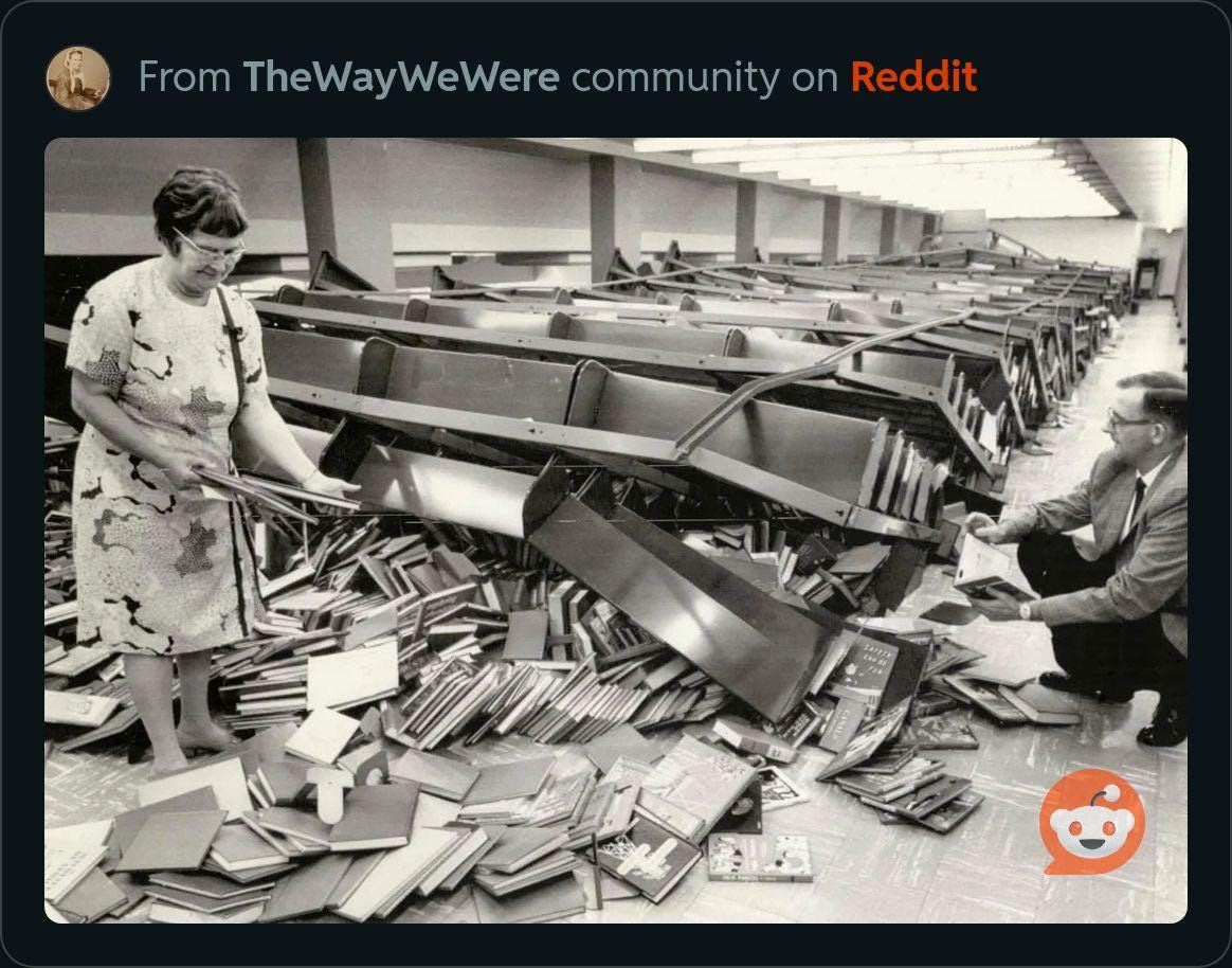 An image taken from The Way We Were Community on reddit shows two people picking up books after bookshelves were knocked over.r/CatastrophicFailure - Library bookshelves fall like dominoes 1971 - aftermath
