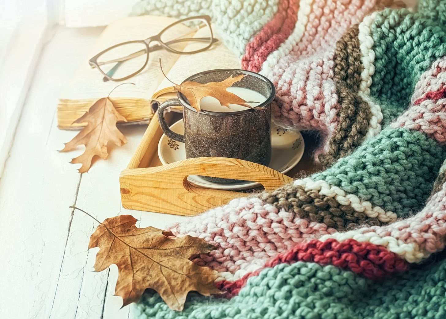 A mug on a wooden tray, surrounded by a colorful knitted blanket and autumn leaves. Glasses rest on an open book nearby.