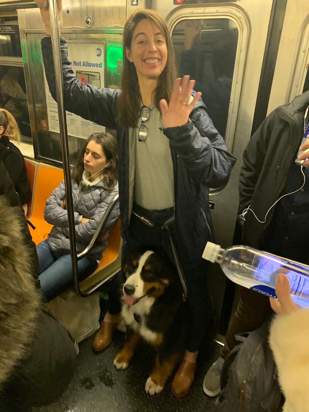 Kate is on a New York City subway, waving to the camera with a forced smile on her face. Her service dog Waffle is beneath her legs. Kate is on a New York City subway, waving to the camera with a forced smile on her face. Her service dog Waffle is beneath her legs.