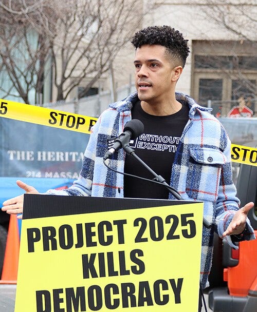 A public speaker stands at a microphone during a protest, holding out his hands in front of a large yellow sign that reads “Project 2025 Kills Democracy.” Yellow caution tape in the background reads “Stop.” The demonstration is taking place outdoors, highlighting opposition to Project 2025 and concern for American democracy. No personal identification is provided. A public speaker stands at a microphone during a protest, holding out his hands in front of a large yellow sign that reads “Project 2025 Kills Democracy.” Yellow caution tape in the background reads “Stop.” The demonstration is taking place outdoors, highlighting opposition to Project 2025 and concern for American democracy. No personal identification is provided.