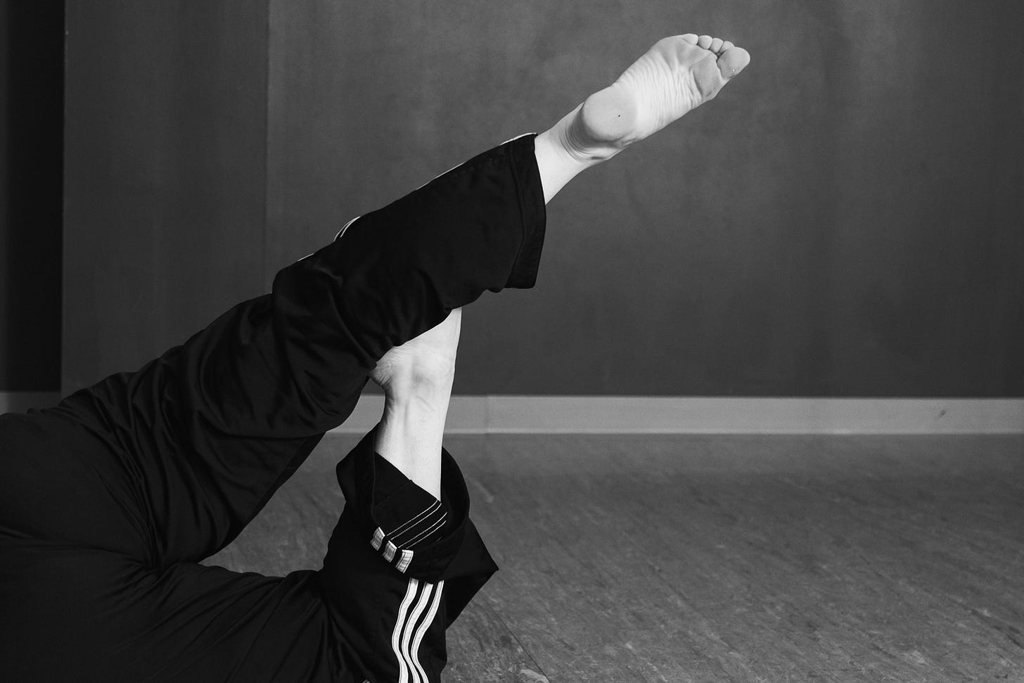 A black and white close-up of a barefoot person from the waist down wearing black pants with a white triple-stripe and cuff detail, who has one leg extended in the air, with a grey textured floor and wall.
