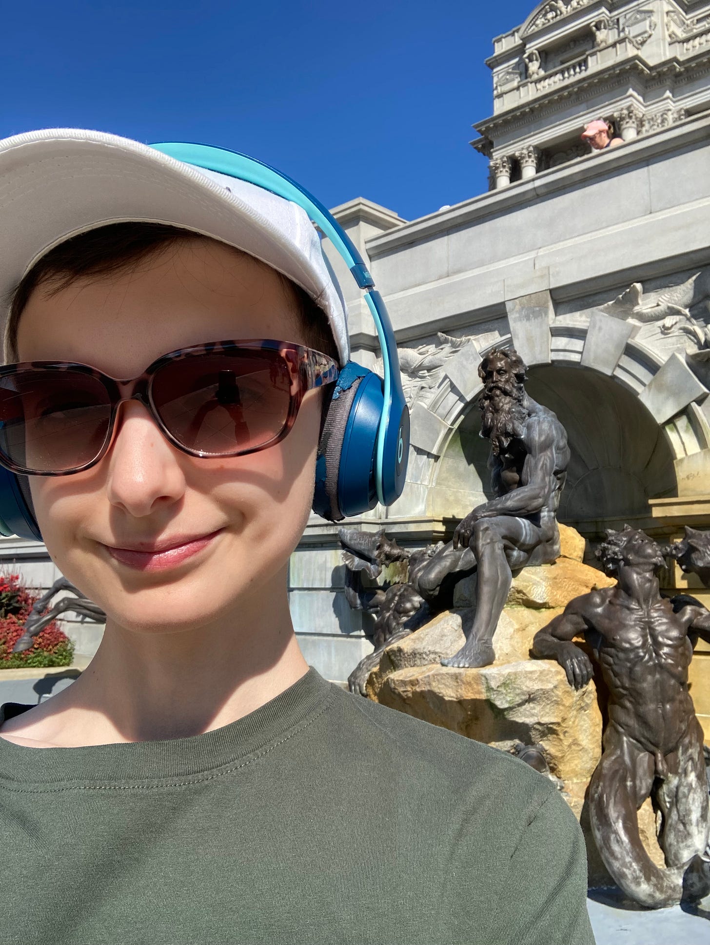 Selfie of Beck wearing headphones, cap, and sunglasses outside the fountain at the Library of congress. Photobombed by the weird naked man statues in the background