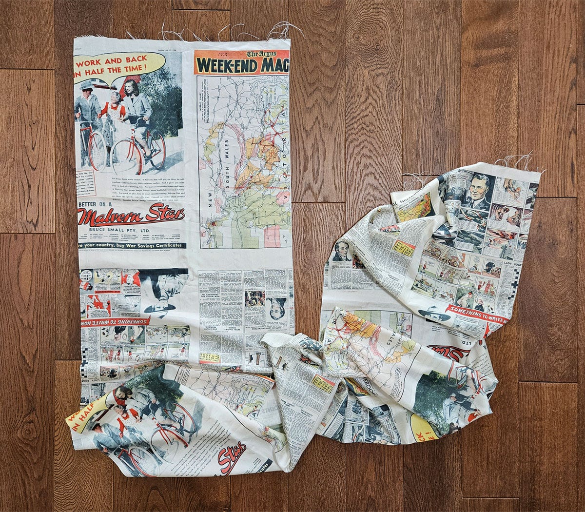 A long piece of silk printed with newspaper pages, laid out on a wooden floor