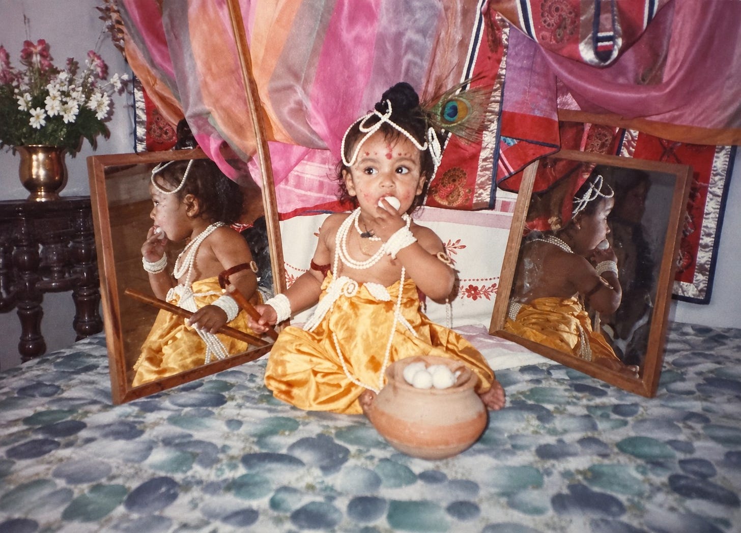 A toddler dressed as baby Krishna, peacock feather in his hair, eating butter from a clay pot, between two mirrors A toddler dressed as baby Krishna, peacock feather in his hair, eating butter from a clay pot, between two mirrors