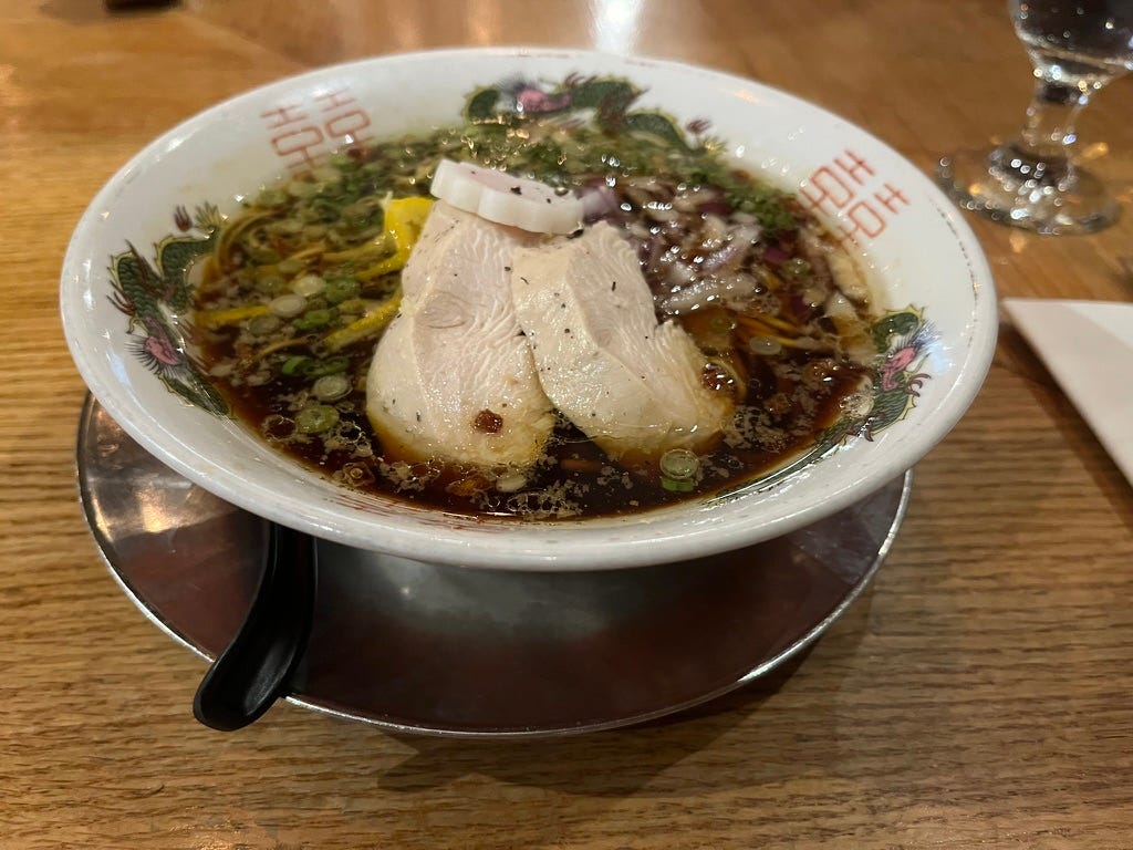 a bowl of clear dark brown soup with green herbs, chopped red onions, and pieces of a yellow citrus fruit, topped with slices of cooked chicken and a naruto