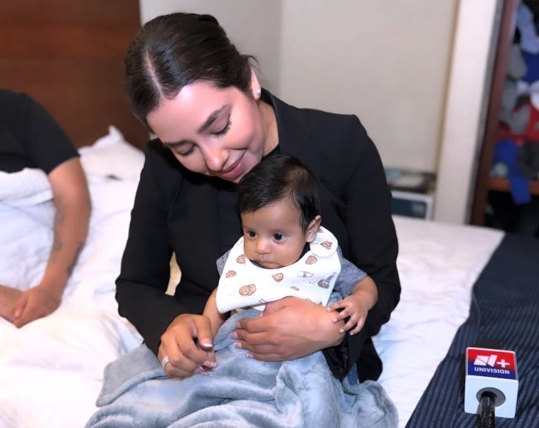 Video screenshot of reporter Lidia Terrazas holding baby Juan Nicolás while sitting at the edge of a hotel bed. His mother is partially in frame at the left of the photo. Terrazas smiles as the looks at the baby, who is wide-eyed and wearing a bib, the tiny fingers of his left hand rest on Terrazas's wrist. Her hand-held wireless microphone, with the Univision logo, sits on the bed next to her.