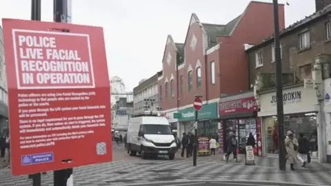 BBC A view of Croydon High Street in February 2024 when the Met deployed live facial recognition (LFR) vans. A red sign warning people that LFR is being used is tied to a lamppost in the foreground, while a white van that has cameras mounted to its roof can be seen in the background as people mill around it and past shops on the street