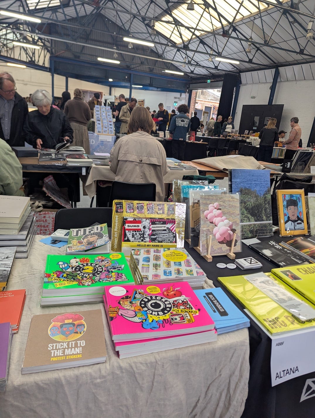 A colourful spread of books on a table at Peckham 24, with multiple Stickerbomb and SOI Books titles visible including Stick It To The Man and Stickerbomb World, with visitors browsing in the background.