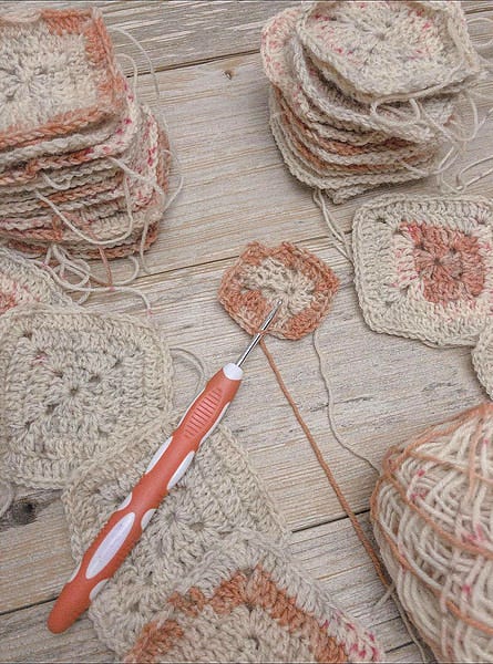 An overhead view of neatly stacked crochet squares in varying stages of completion for a Battenberg blanket. The squares feature warm neutral and peach tones. In the center lies a crochet hook with an ergonomic orange handle, placed next to an individual square partially completed, emphasizing the crafting process. Loose yarn ends are visible, adding to the handmade, work-in-progress ambiance of the image, set against a rustic wooden background. An overhead view of neatly stacked crochet squares in varying stages of completion for a Battenberg blanket. The squares feature warm neutral and peach tones. In the center lies a crochet hook with an ergonomic orange handle, placed next to an individual square partially completed, emphasizing the crafting process. Loose yarn ends are visible, adding to the handmade, work-in-progress ambiance of the image, set against a rustic wooden background.