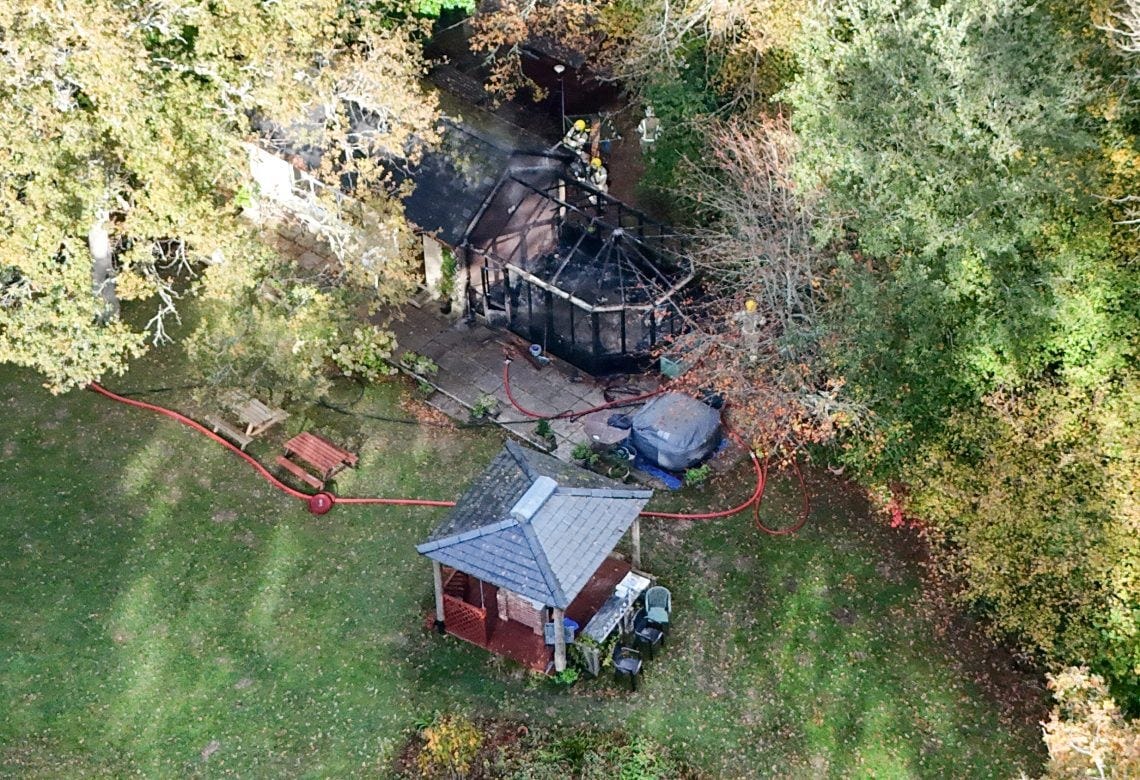Aerial view of a fire-damaged building surrounded by trees at the Valerian Sun Club on the Isle of Wight. The conservatory’s frame is blackened and collapsed, with fire hoses stretched across a grassy clearing and firefighters working near the structure. Aerial view of a fire-damaged building surrounded by trees at the Valerian Sun Club on the Isle of Wight. The conservatory’s frame is blackened and collapsed, with fire hoses stretched across a grassy clearing and firefighters working near the structure.