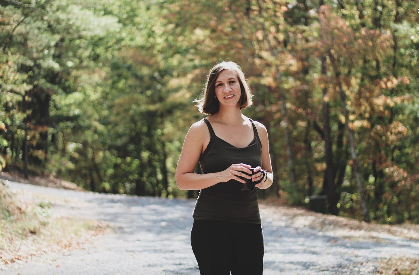 A white, brunette woman in an olive tank top and black pants holds a black coffee mug on a trail in the woods