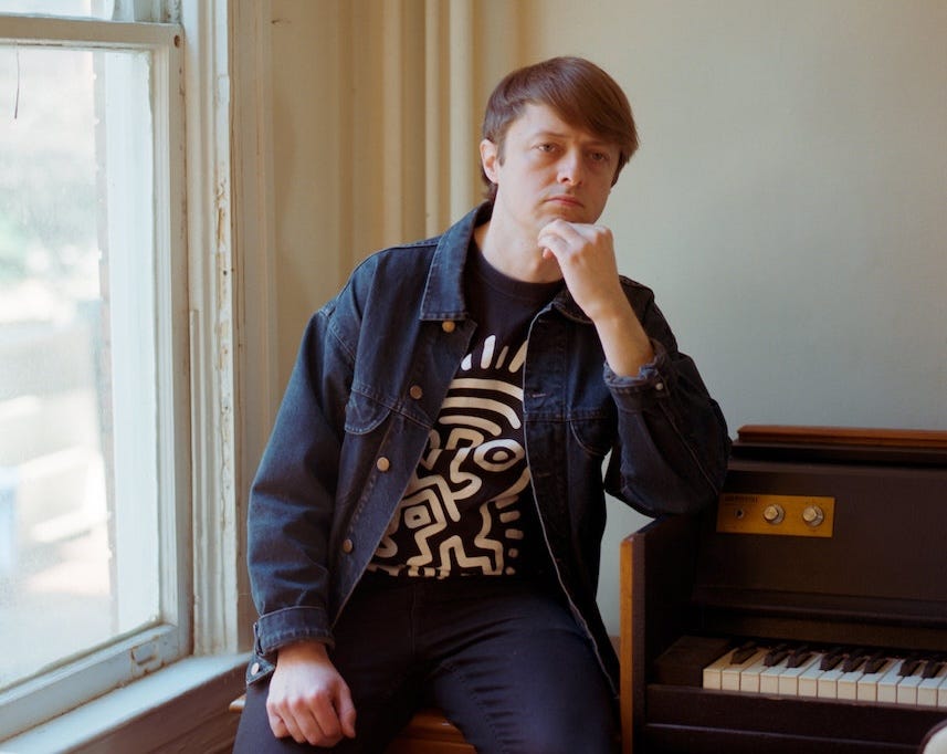 man in denim jacket with hand on chin and arm on piano next to window