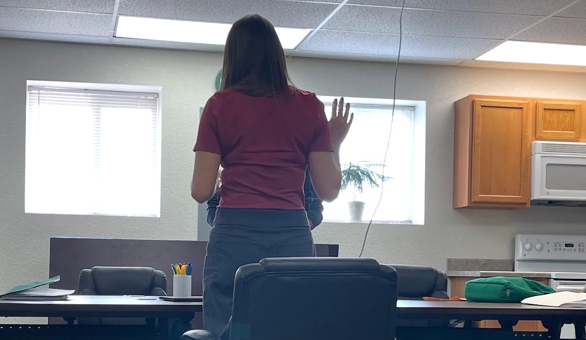 woman in a red tshirt and blue pants with her back to the camera raises her right hand at a small board table and reads from a piece of paper held in her left hand woman in a red tshirt and blue pants with her back to the camera raises her right hand at a small board table and reads from a piece of paper held in her left hand