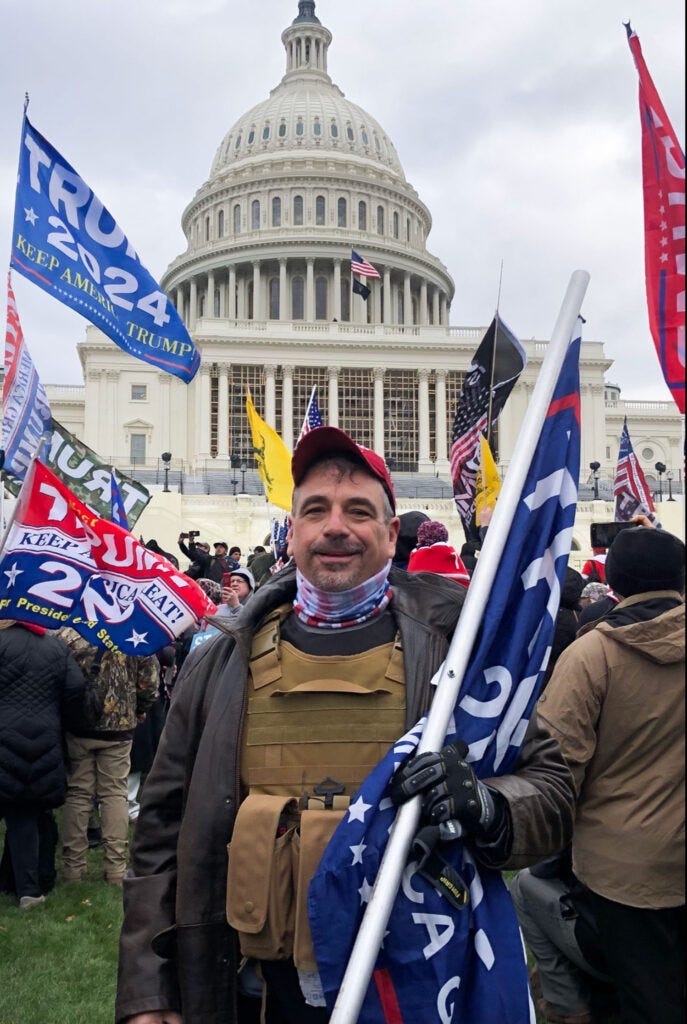 Man in foreground amid a large crowd wearing a bullet-proof vest and holding a flag pole with the US Capitol building in the background. Man in foreground amid a large crowd wearing a bullet-proof vest and holding a flag pole with the US Capitol building in the background.