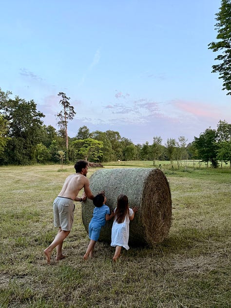 Golden evening light on the château grounds: a child in a white nightgown walking toward the coach house past hay bales; a girl on a stone path with the château behind; a man and two children pushing a large round hay bale.