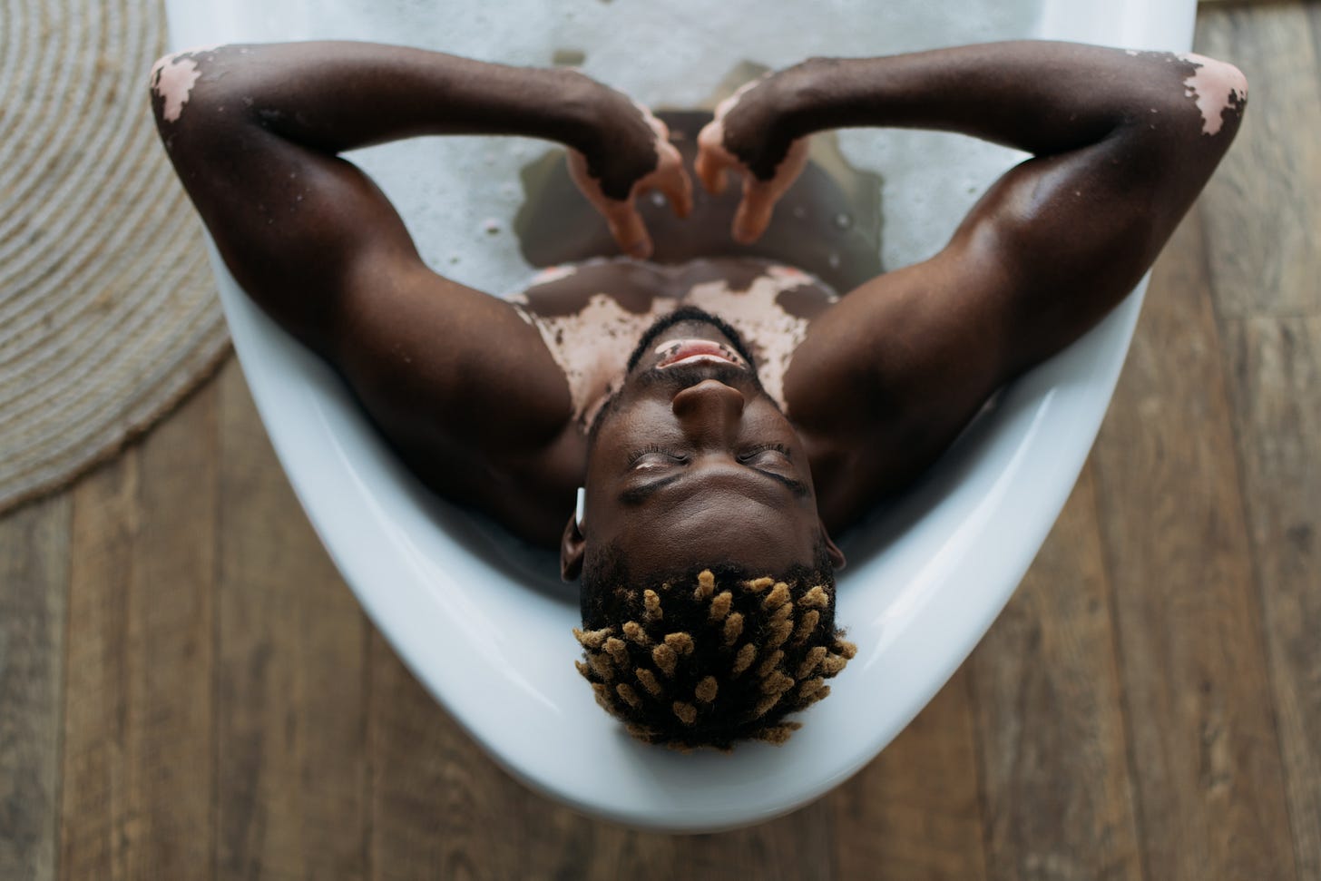 Overhead view of a nude Black man with vitiligo relaxing in a bathtub, eyes closed, arms bent gently upward, and a single wireless earbud in his left ear, listening to music.