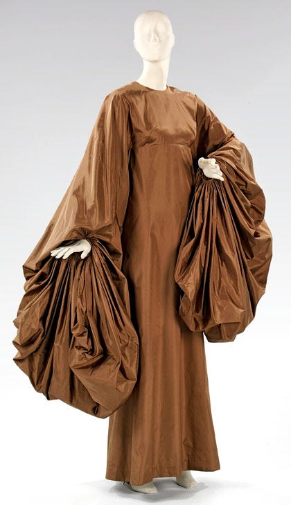 A white mannequin wears an ankle-length tobacco brown dress in crisp silk paper taffeta with oversized, gathered sleeves that drape toward the ground.