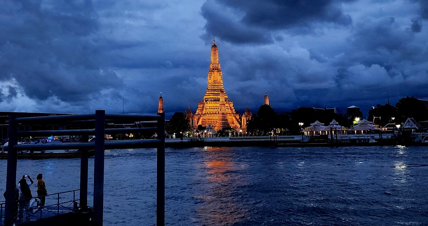 Wat Arun silhouetted at dusk beneath dramatic dark clouds, reflected in the Chao Phraya River in Bangkok, Thailand.