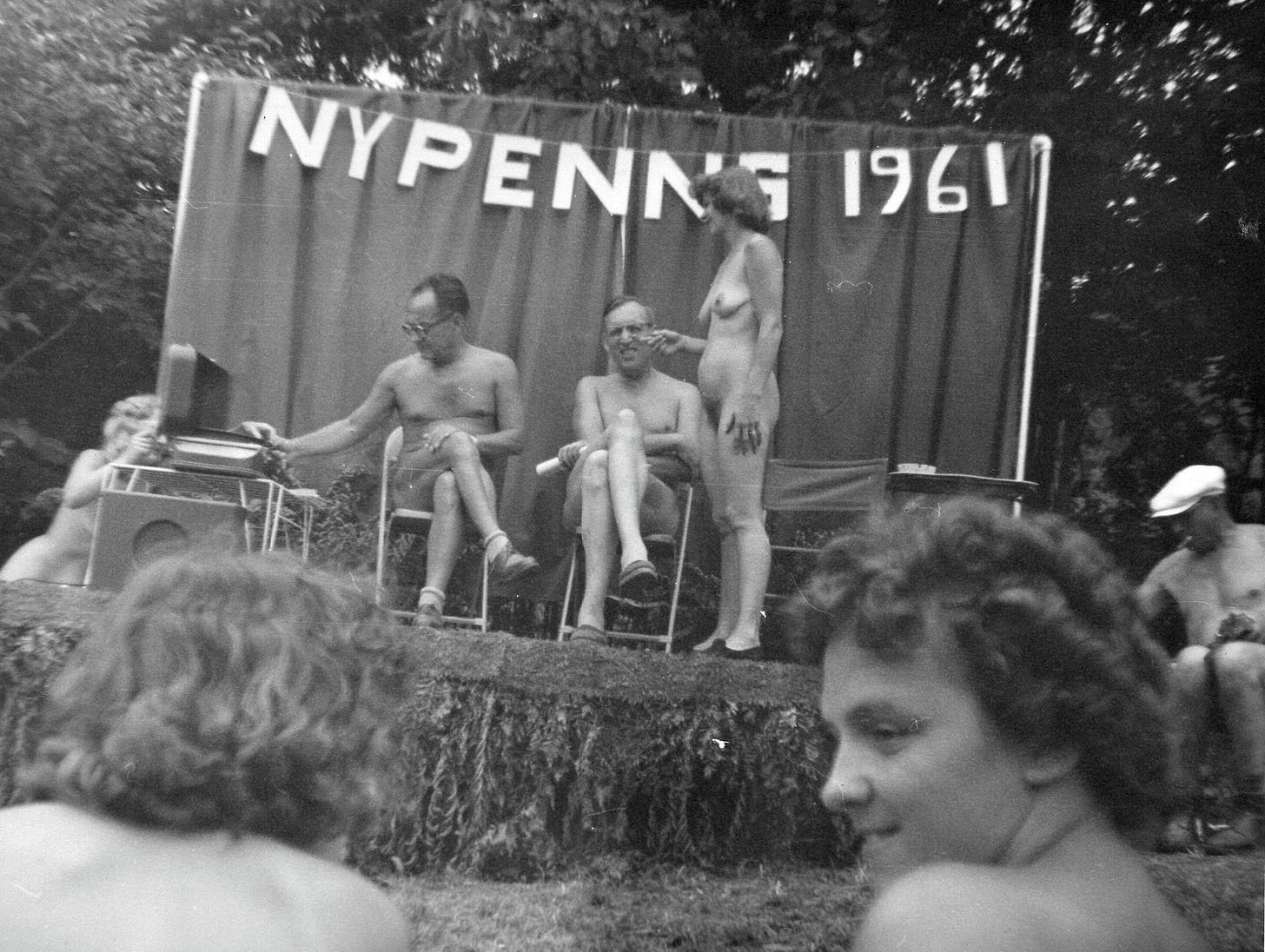 A small nude gathering in front of a banner reading “NYPENNS 1961,” with several adults seated on a platform and others nearby.