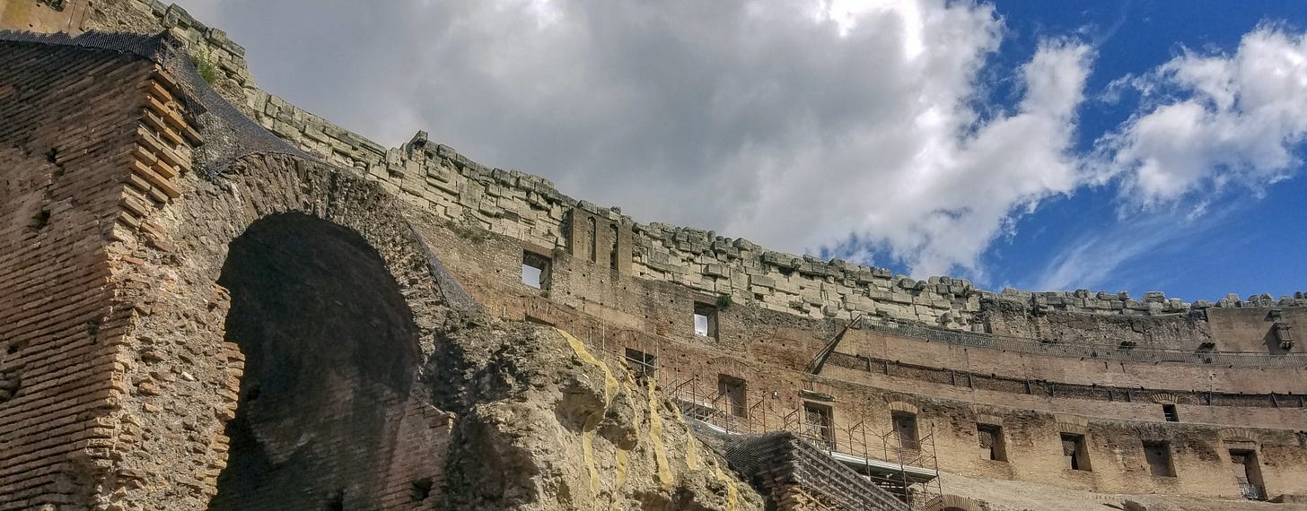 Photograph of the Roman Coliseum, from the inside looking up at the exposed rim, with blue sky and clouds in background.