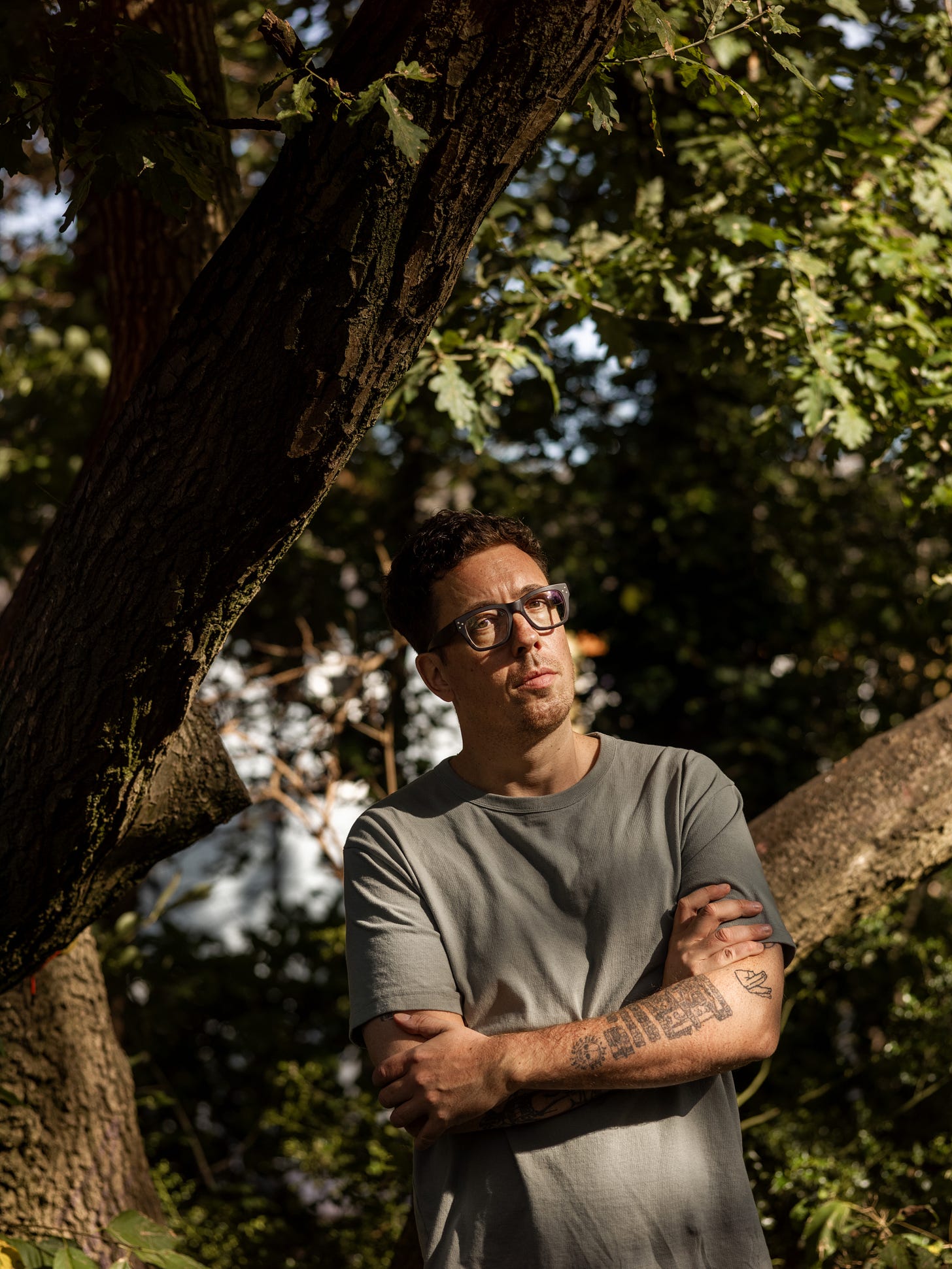 A tall dark haired man with spectacles and tattoos stands in a grey-green tshirt with his arms folded, against a shadowy background of trees and foliage. 