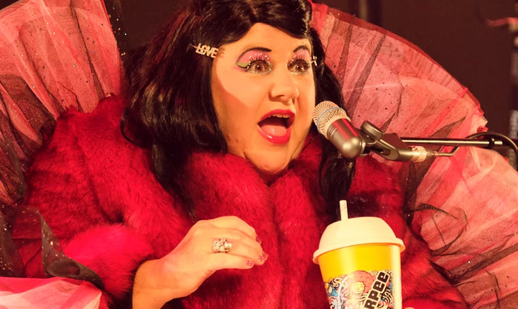 A performer in a vibrant red outfit with pink and black tulle accents speaks into a microphone. They wear glittery makeup, bold lipstick, and a hairpin that spells ‘LOVE’. In one hand, they hold a yellow Slurpee cup.
