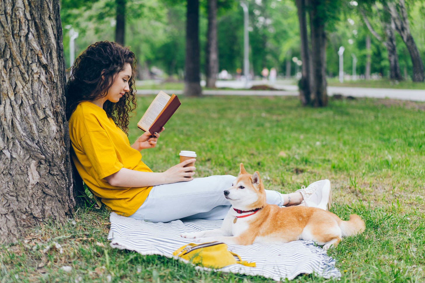 woman leans against tree while reading book, dog laying next to her