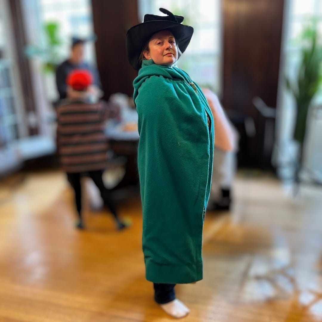 A person dressed in a dramatic black hat and a long green cloak stands confidently in the center of a room, looking directly at the camera. The background is softly blurred, showing other people engaged in activities, adding to the sense of a lively gathering.