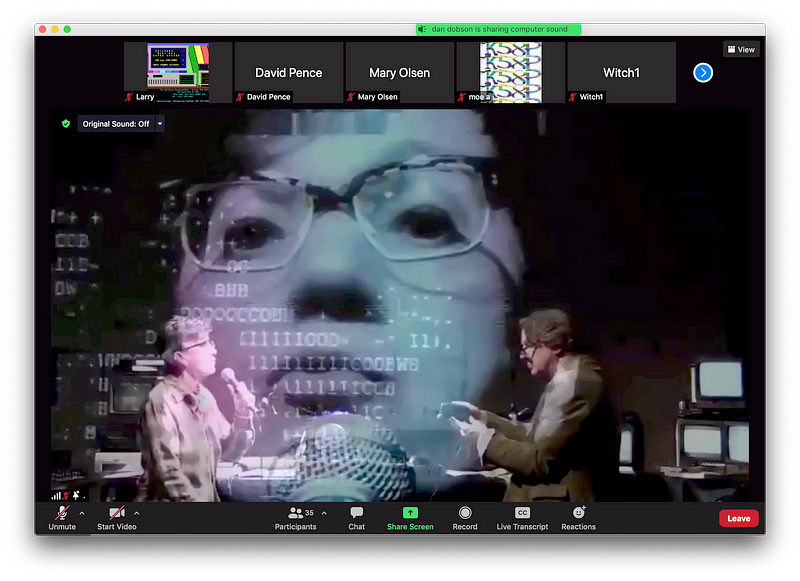 The image is a screenshot of a computer monitor. There’s a border of various instructions (“unmute,” “start video,” etc.). In the center are two people, facing each other. They’re in a darkened room, with electronic equipment surrounding them. One person is holding a microphone and the other cradles a small video camera. A projected image of the person with the microphone is magnified and superimposed, creating a surreal collage of imagery.