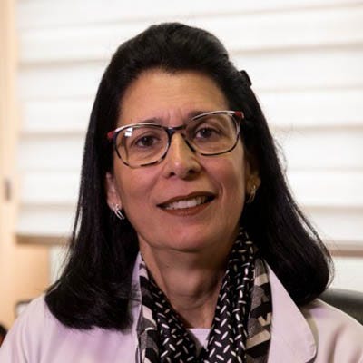 A imagem mostra uma mulher enquadrada do peito para cima olhando diretamente para a câmera. Ela tem pele clara, cabelos escuros, lisos e de comprimento médio, penteados para trás. Usa óculos de armação retangular e esboça um sorriso. Ela veste um jaleco branco, e, por baixo, há um lenço ou echarpe escuro com padrão claro, enrolado no pescoço. Ao fundo, o ambiente parece interno e bem iluminado, com uma parede clara e uma janela com persianas horizontais fechadas.