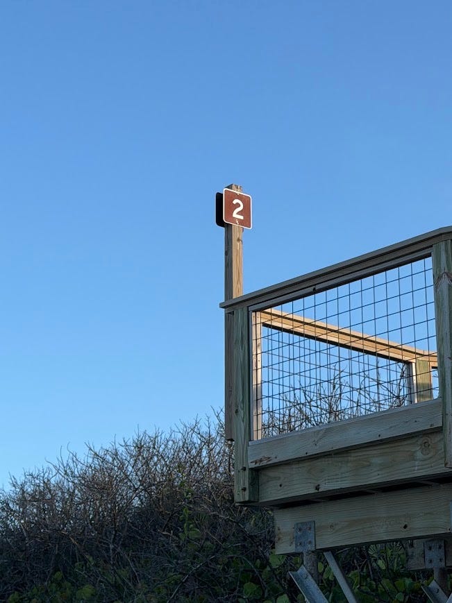 Photo of a small brown sign with the number two on it on a wooden beach walkway against a blue sky.