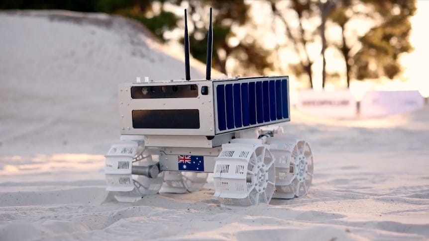 A small square robot has four white wheels, it wheels through white sand