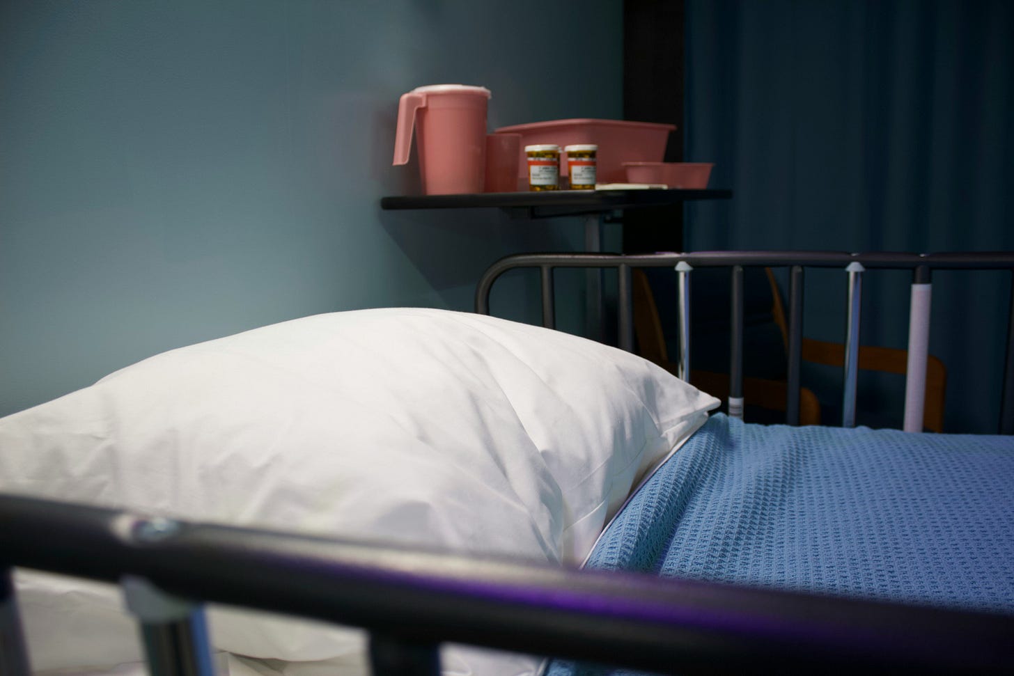 An empty hospital bed with blue sheets and pink plastic pitcher on a shelf in the background. 