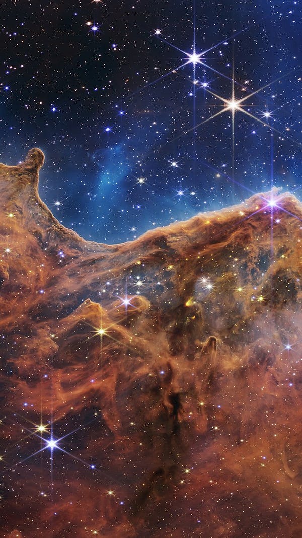 The image is divided horizontally by an undulating line between a cloudscape forming a nebula along the bottom portion and a comparatively clear upper portion. Speckled across both portions is a starfield, showing innumerable stars of many sizes. The smallest of these are small, distant, and faint points of light. The largest of these appear larger, closer, brighter, and more fully resolved with 8-point diffraction spikes. The upper portion of the image is blueish, and has wispy translucent cloud-like streaks rising from the nebula below. The orangish cloudy formation in the bottom half varies in density and ranges from translucent to opaque. The stars vary in color, the majority of which, have a blue or orange hue. The cloud-like structure of the nebula contains ridges, peaks, and valleys – an appearance very similar to a mountain range. Three long diffraction spikes from the top right edge of the image suggest the presence of a large star just out of view. See less