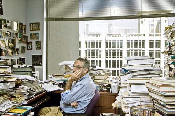 A man cups his face with his right hand while sitting in an office overflowing with papers and books. A man cups his face with his right hand while sitting in an office overflowing with papers and books.