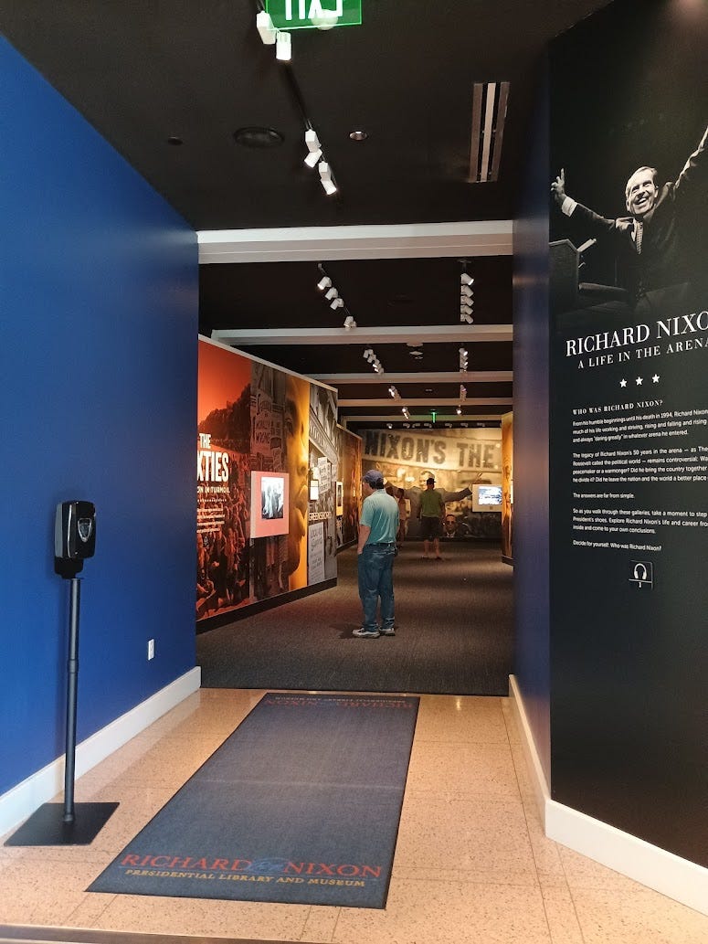 Exhibits in hallway, with only "The Sixties" visible, and Nixon with outstretched arms and "Nixon's the One" sign visible at end.