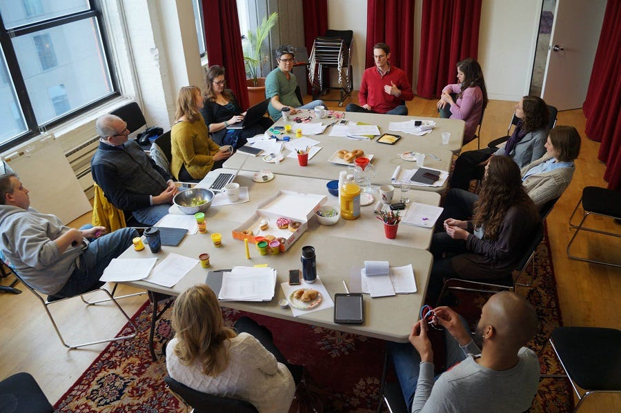 A group of people in a studio sit around a big table full of notebooks, loose paper, pencils, and a box of donuts A group of people in a studio sit around a big table full of notebooks, loose paper, pencils, and a box of donuts